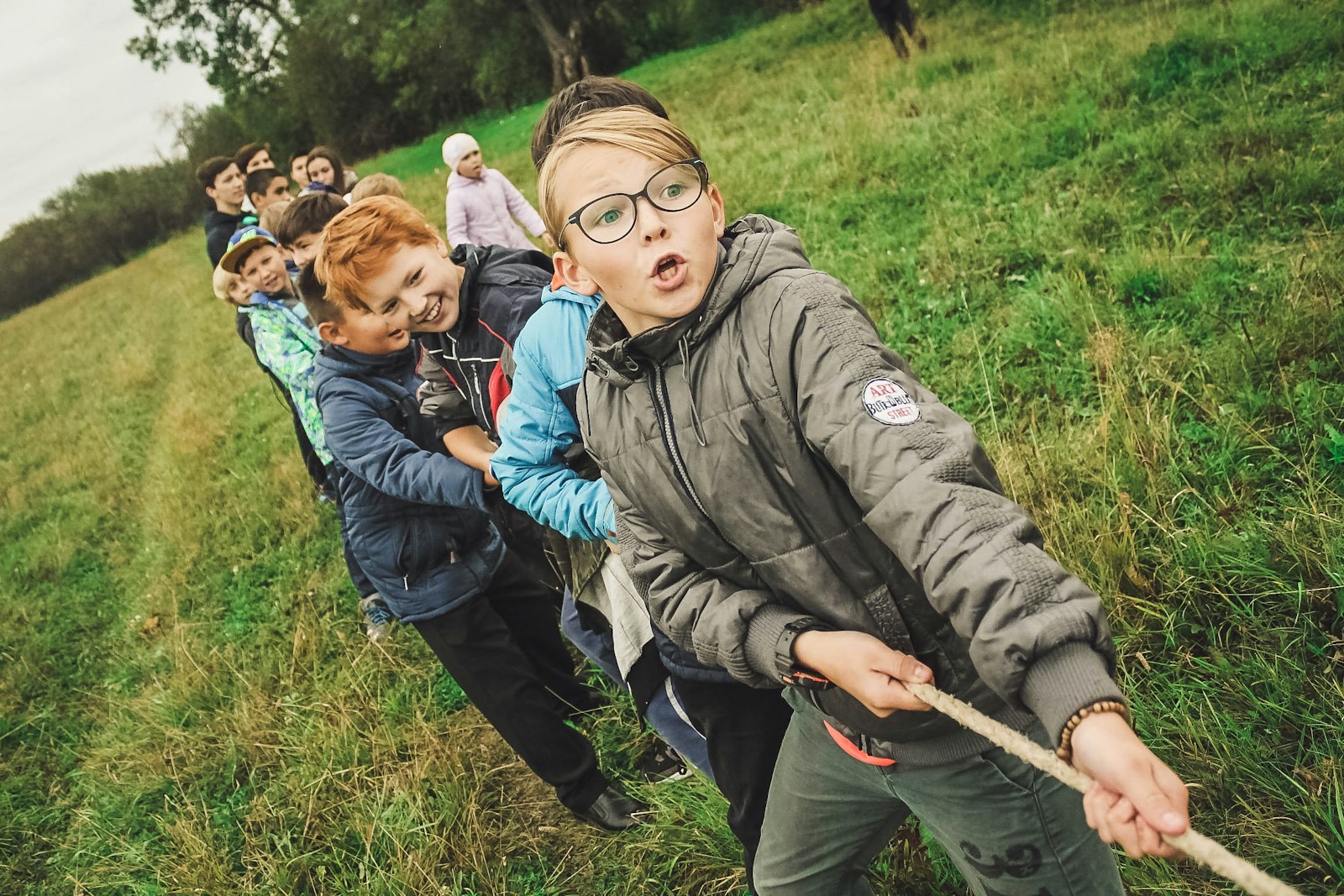 Een rij kinderen trekt lachend en met volle kracht aan een touw op een grasveld